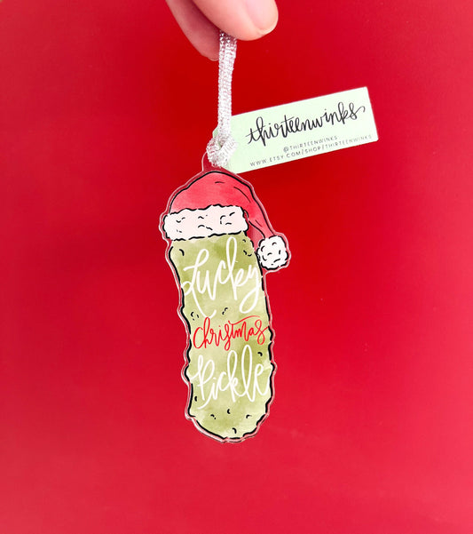 adorable watercolor pickle with a Santa hat. It says Lucky Christmas Pickle in calligraphy on the ornament.